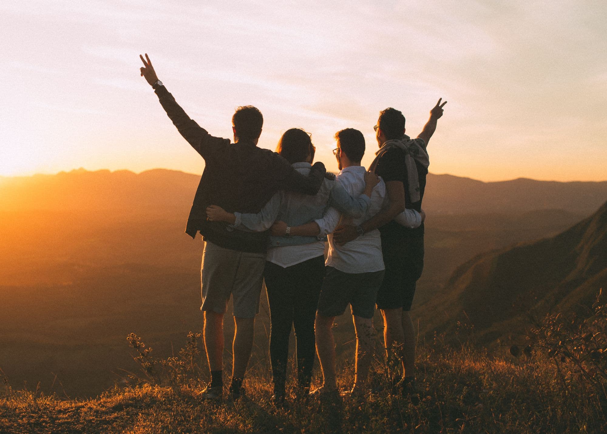 Four friends arm in arm on a hilltop at sunset, raising their hands toward the view